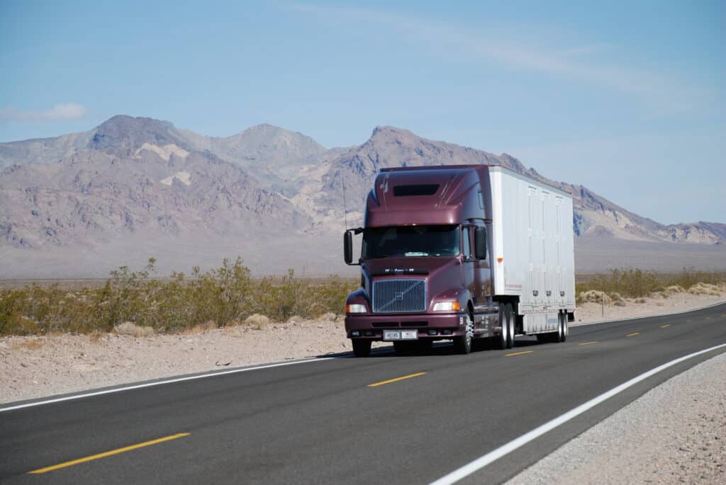A maroon semi-truck travels along a remote desert highway with rugged mountains in the background.