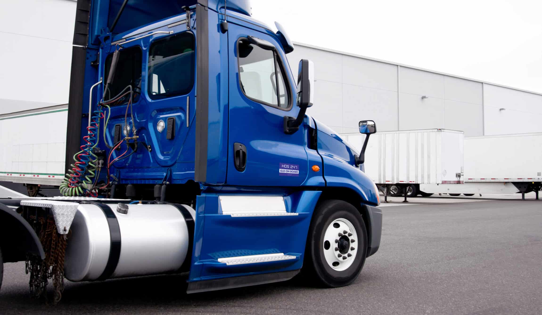 A blue semi-truck cab with an HOS247 logo is parked in an industrial lot, with trailers and a warehouse building visible in the background