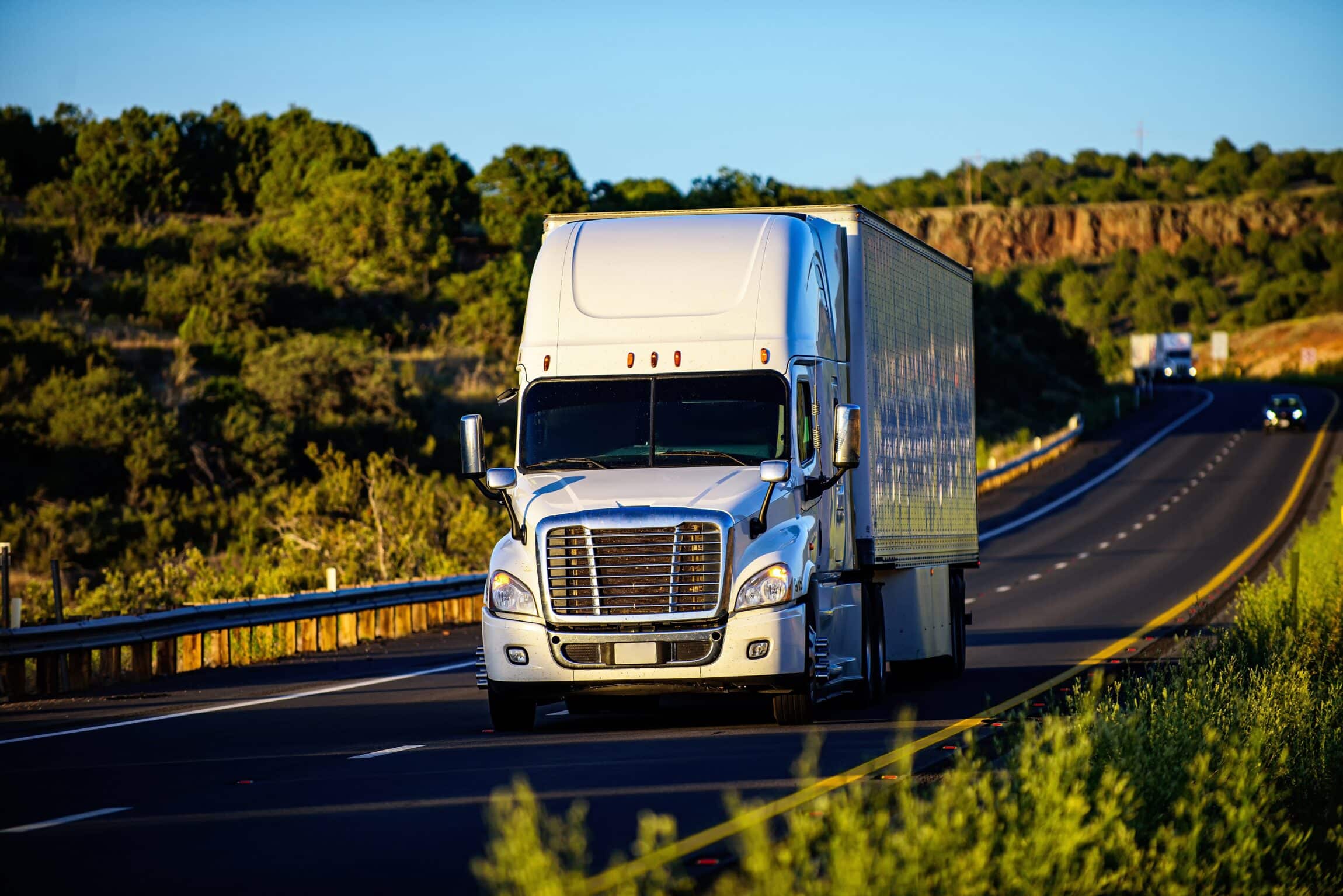 A white semi-truck drives along a scenic highway under a clear blue sky.