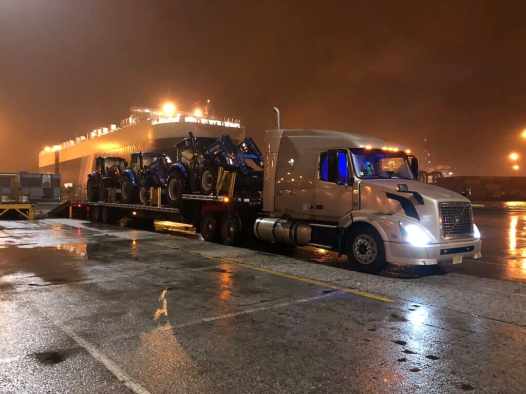 Volvo flatbed semi-truck loaded with blue tractors parked at a well-lit shipping port at night.