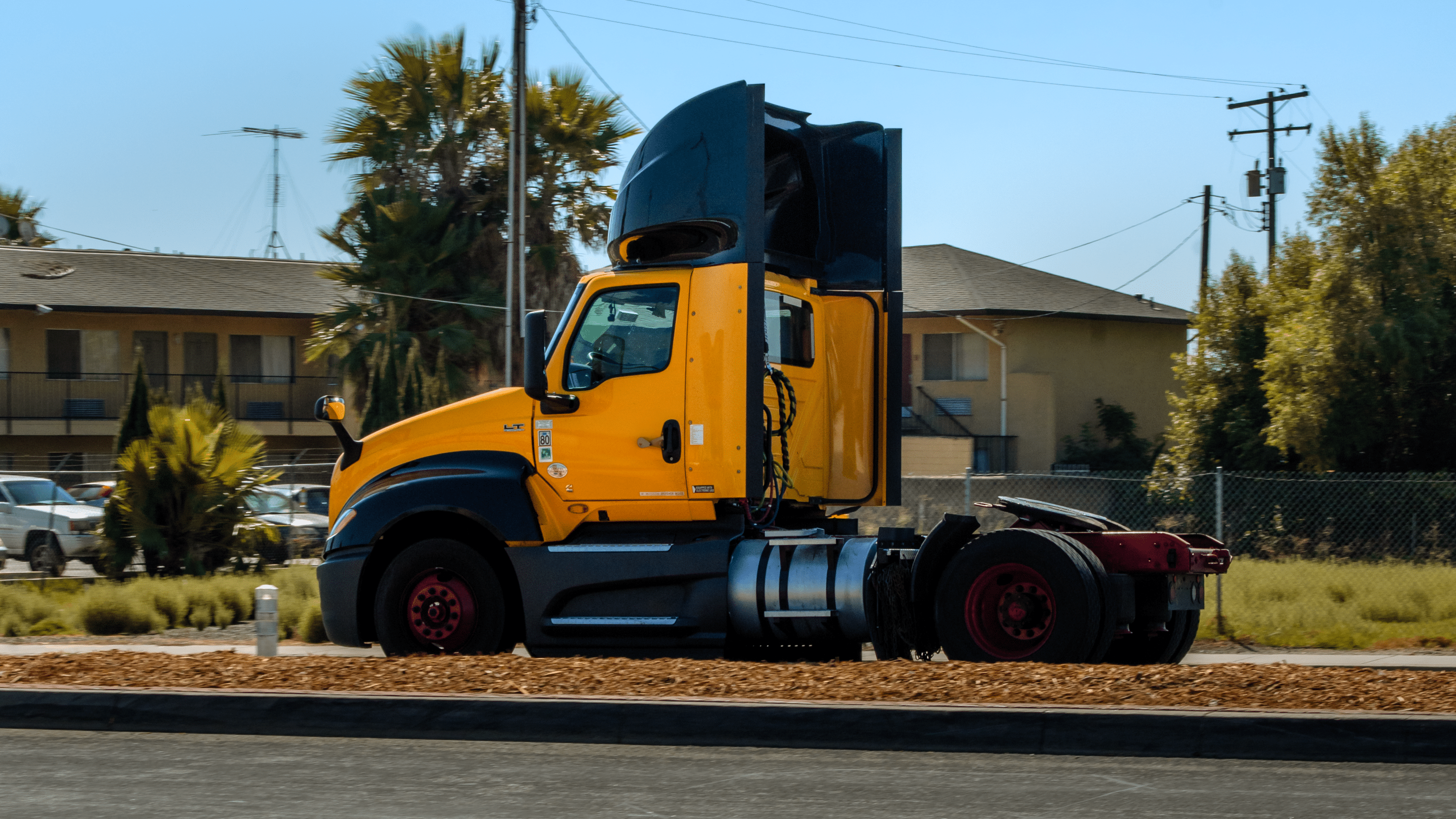 Yellow semi-truck tractor driving on a roadway near residential buildings and palm trees