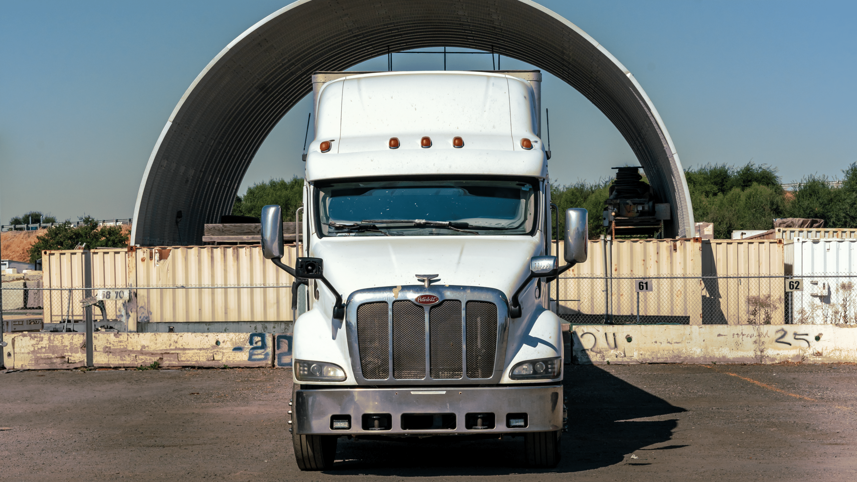 Front view of a white semi-truck parked in an industrial yard with shipping containers in the background