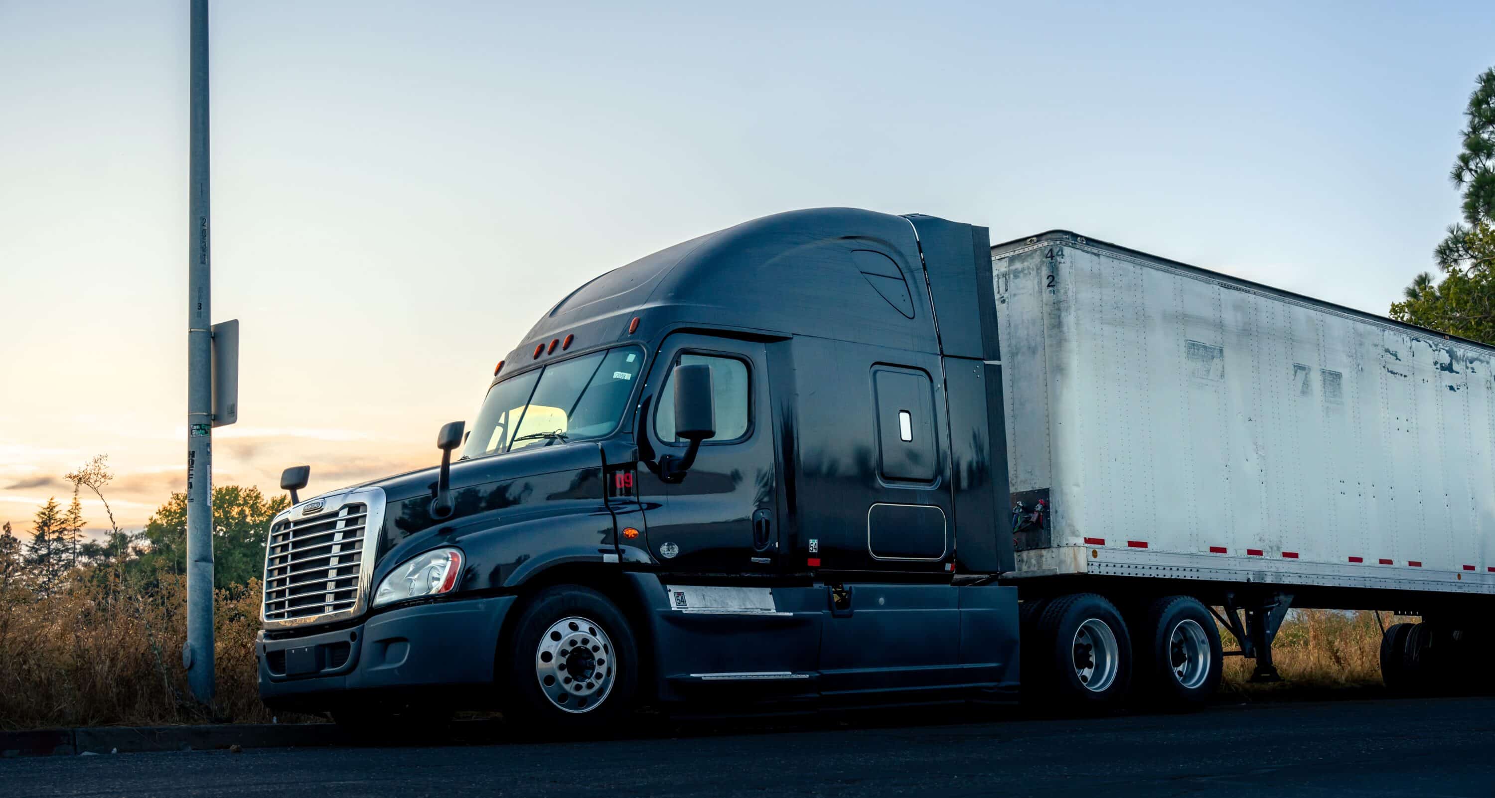 Black semi-truck with box trailer parked beside a roadside at sunset