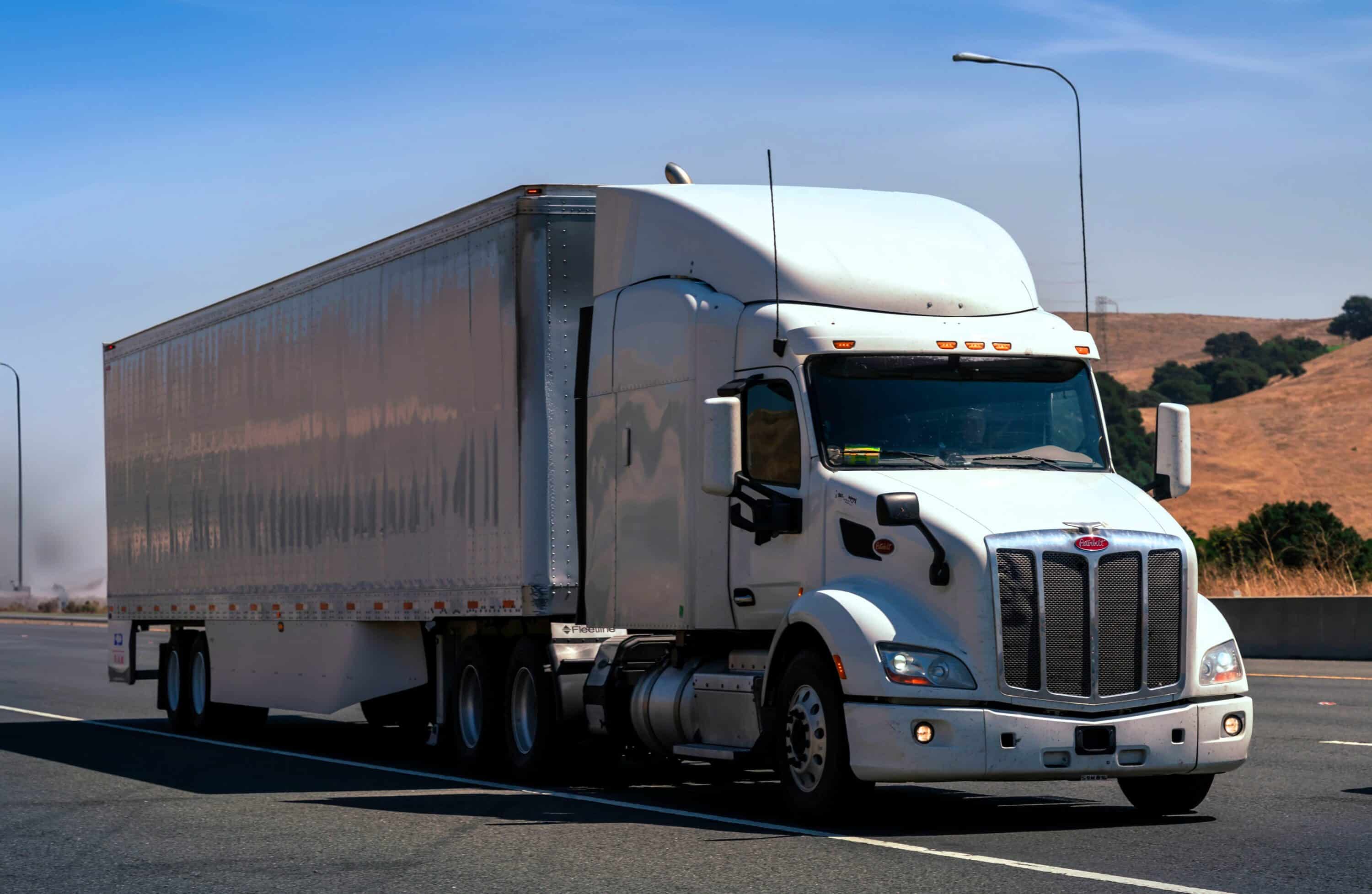 White semi-truck hauling a box trailer on a highway through a dry hilly landscape