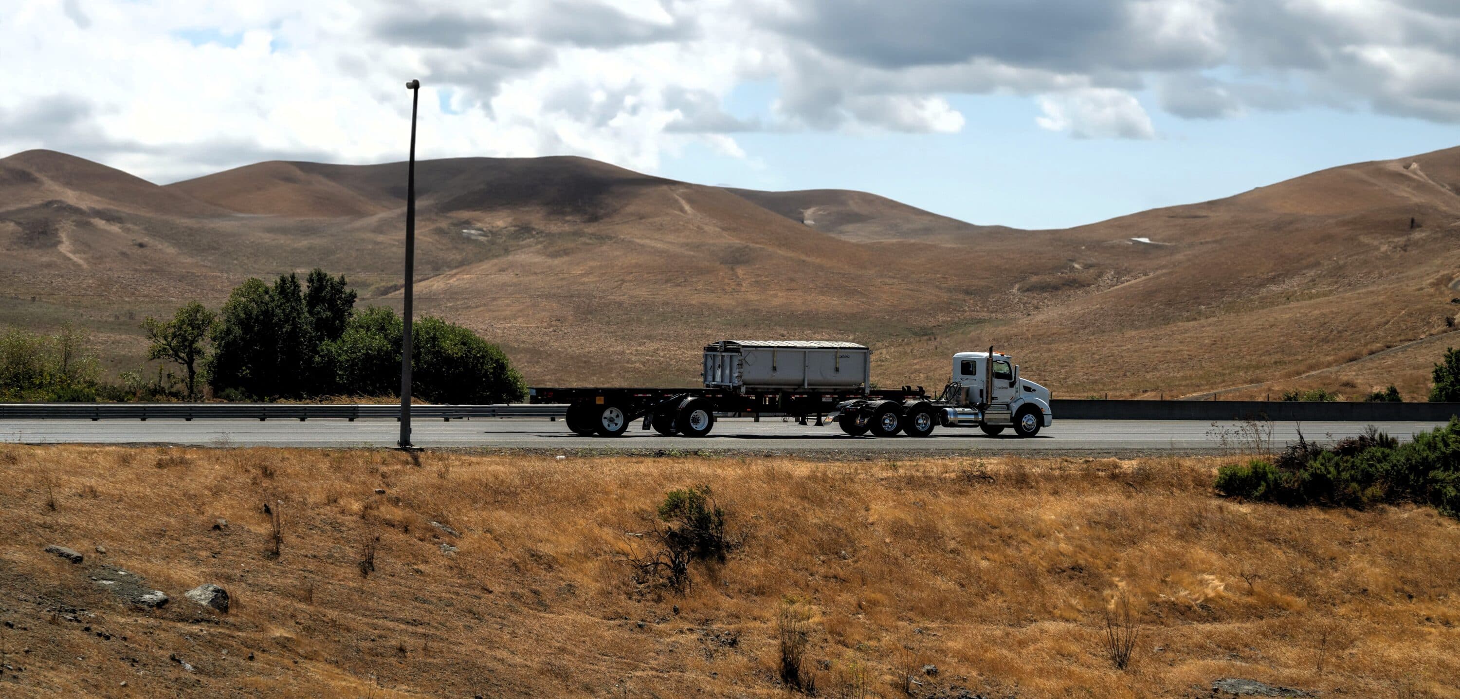 Semi-truck pulling a flatbed trailer along a highway surrounded by rolling hills and open terrain