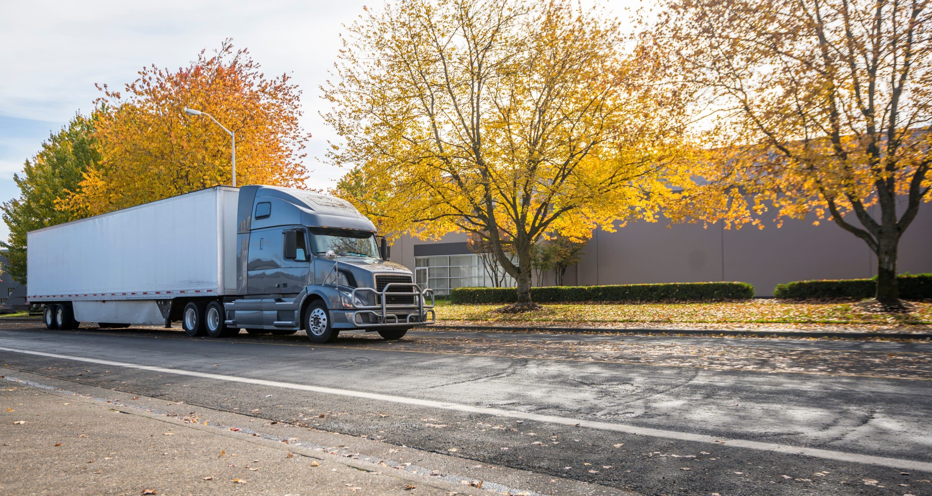 Gray semi-truck with box trailer parked on a street lined with autumn trees