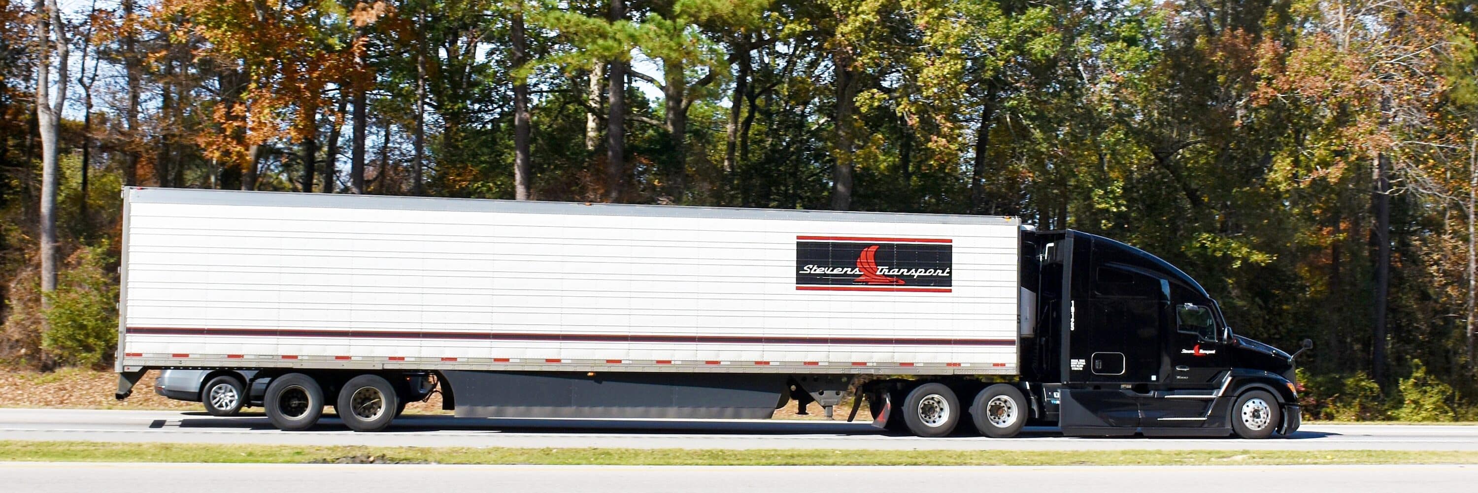 Black semi-truck hauling a white trailer alongside a roadway bordered by autumn foliage