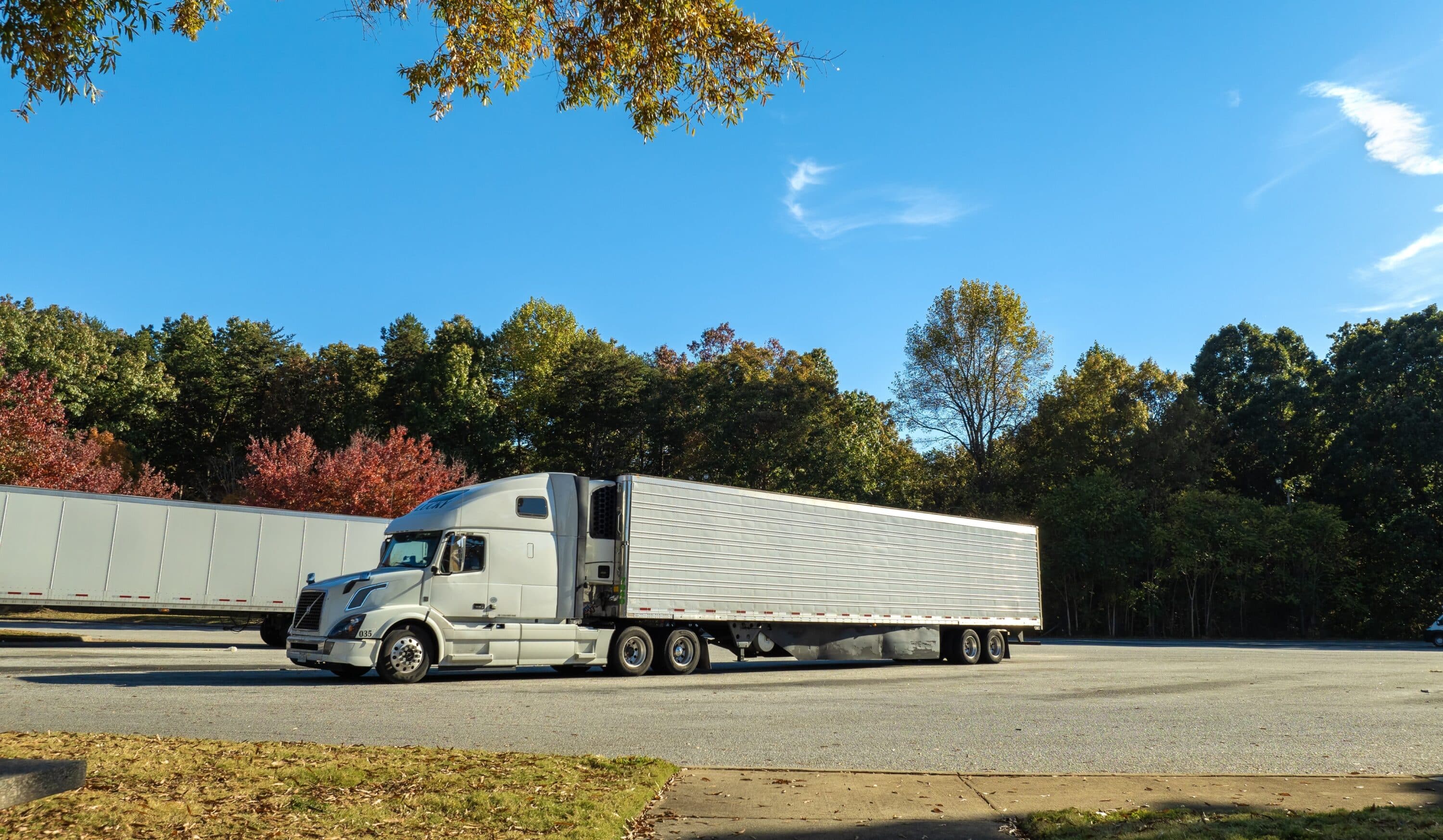 White semi-truck with refrigerated trailer parked in a large lot near a wooded area