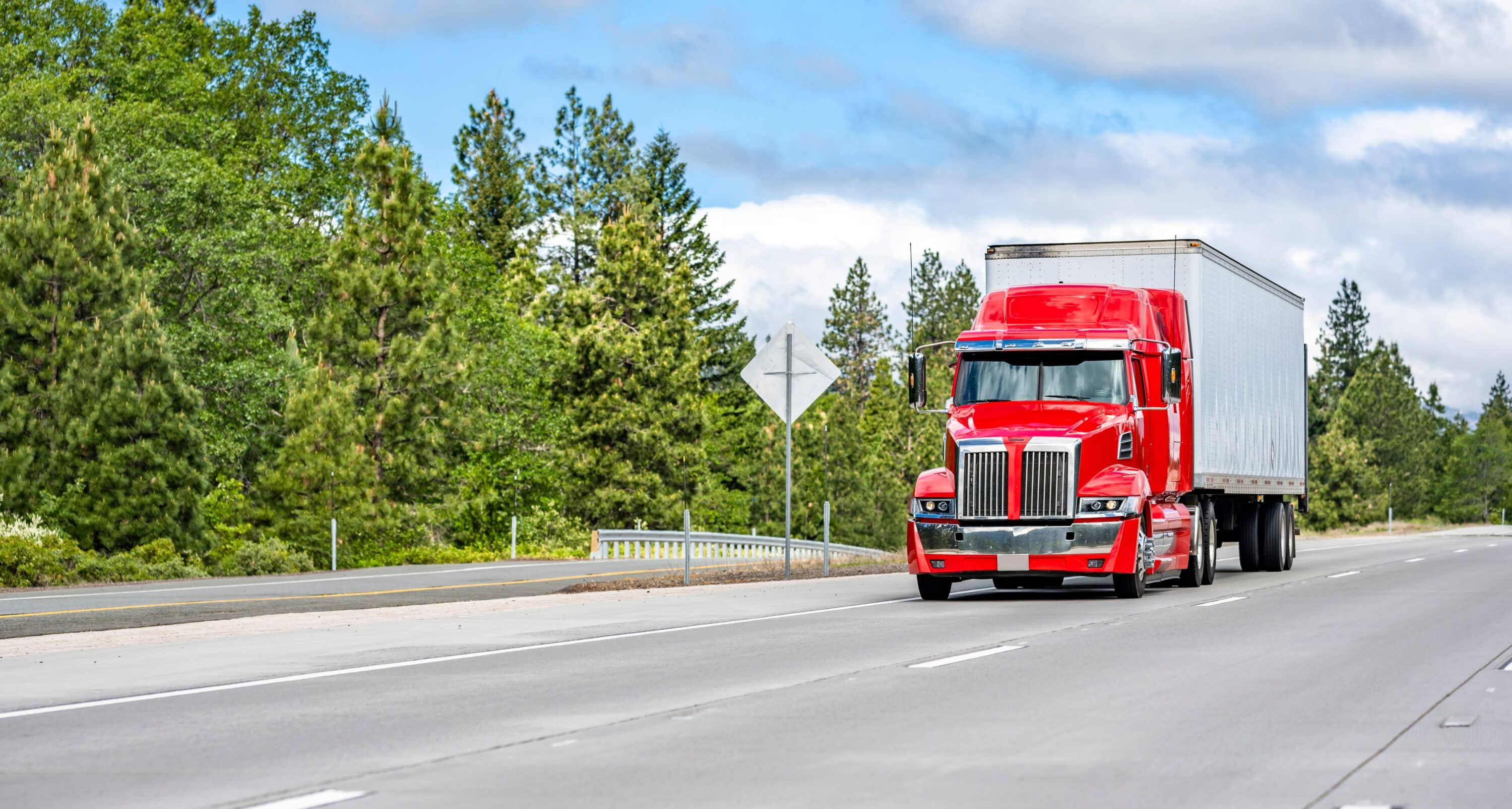 Red semi-truck hauling a box trailer on a highway bordered by trees