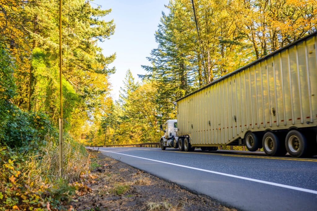 Semi-truck hauling a long trailer on a two-lane road surrounded by trees and autumn foliage
