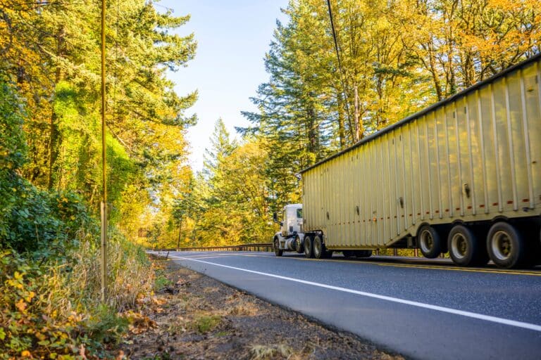 Semi-truck hauling a long trailer on a two-lane road surrounded by trees and autumn foliage