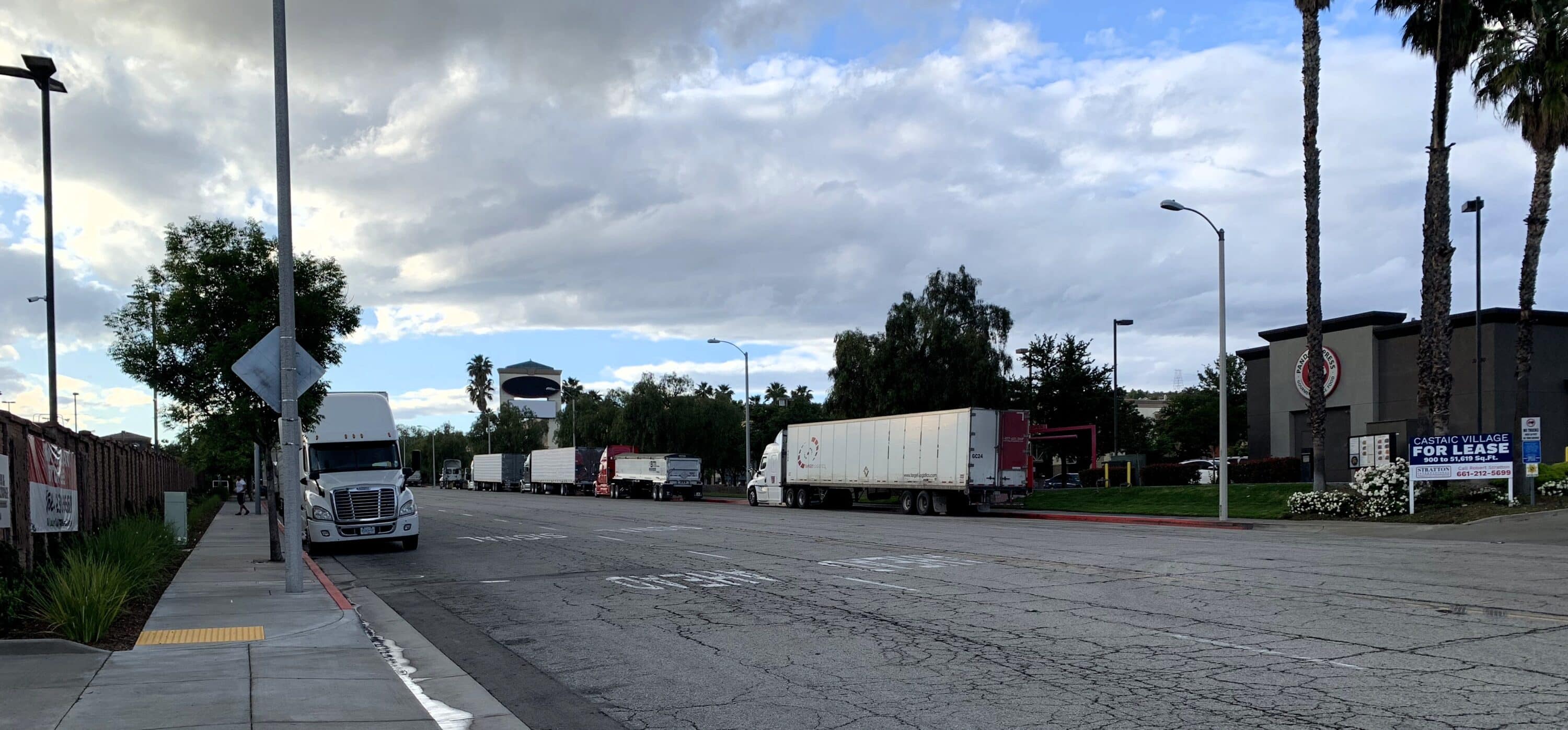 Semi-trucks and trailers parked along a commercial street near business buildings under a cloudy sky