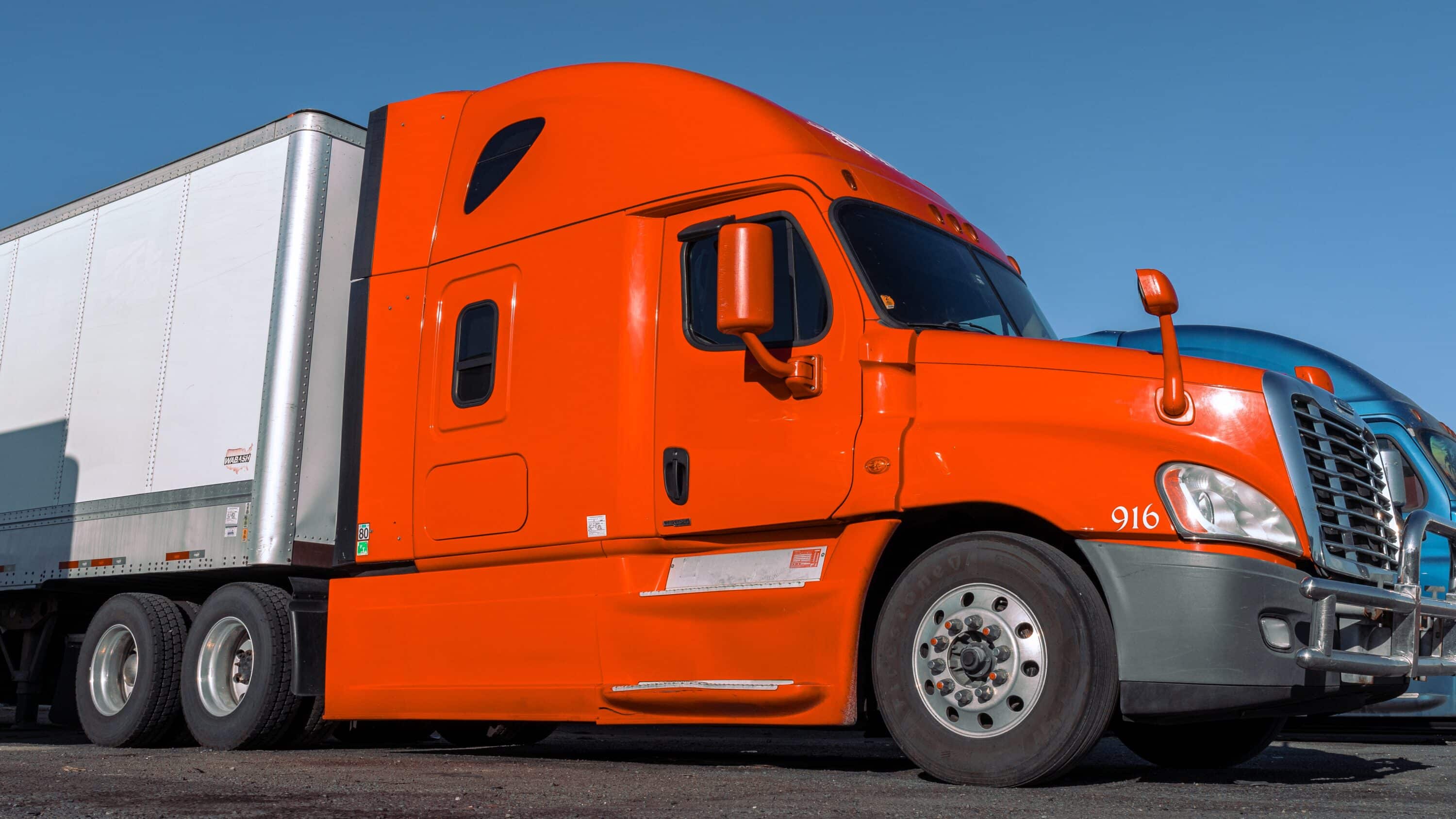 Orange semi-truck with box trailer parked in a commercial truck lot under a clear sky