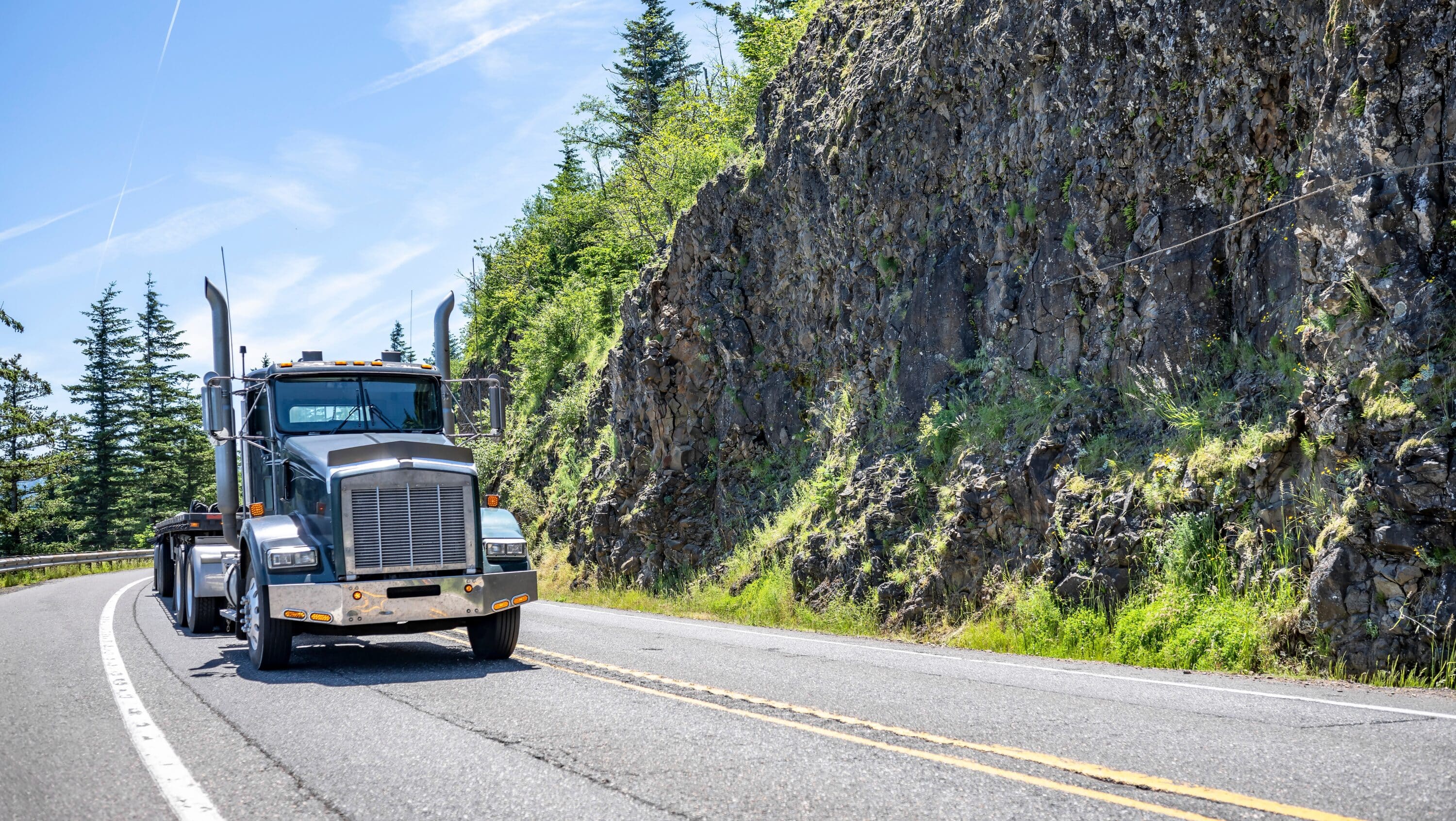 Gray semi-truck driving on a mountain road beside a rocky hillside