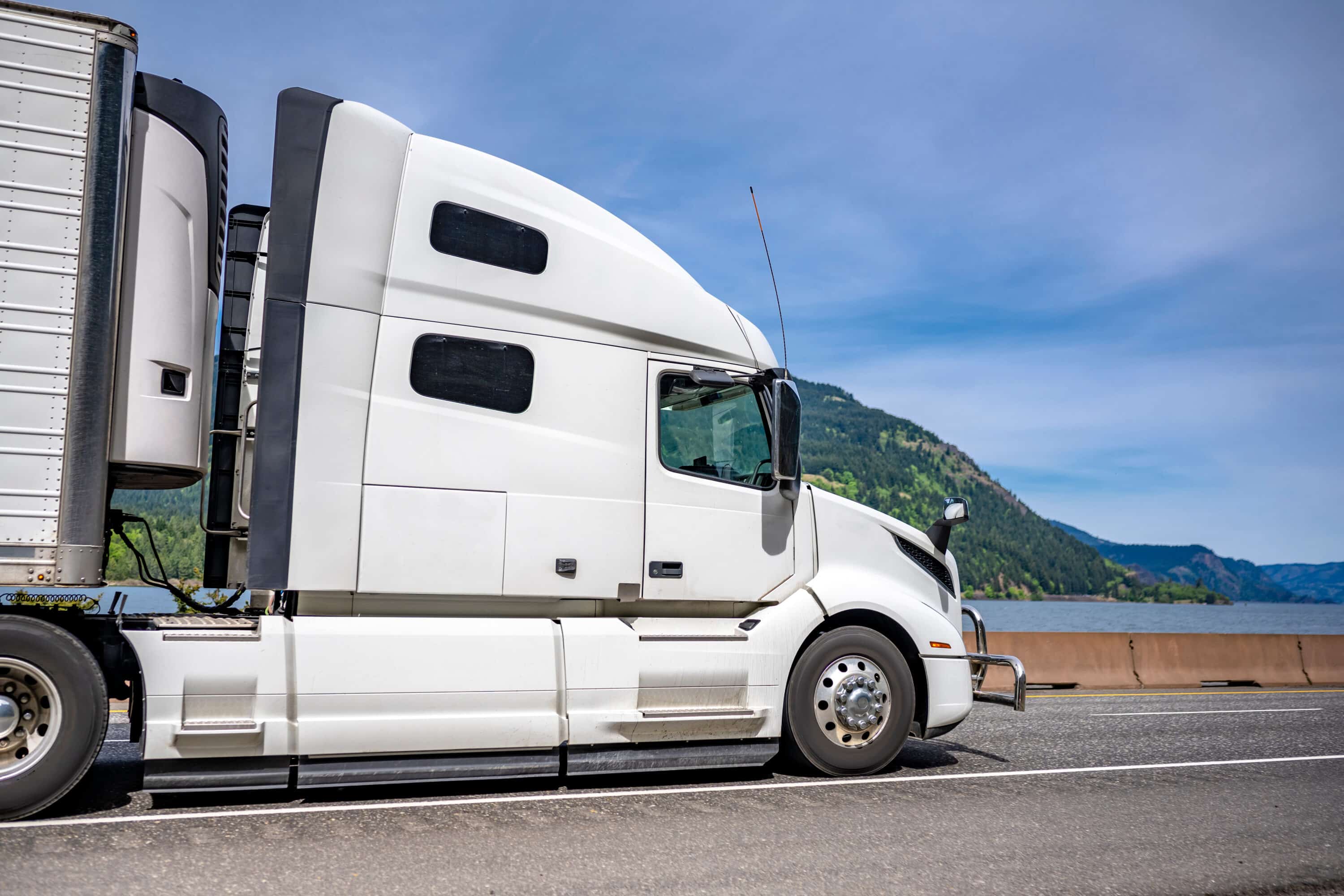 White semi-truck hauling a trailer on a highway with mountains and water in the background